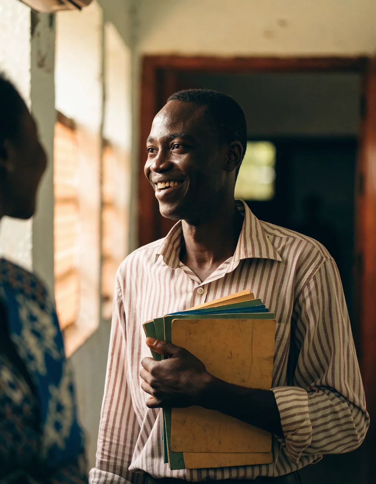 A teacher in an Accra school corridor holding a stack of exercise books